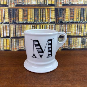May include: White ceramic mug with a black and white striped letter "M" on the front. The mug has a rounded base and a handle. The background features a bookshelf.