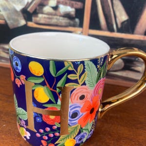 May include: A ceramic mug with a gold handle and the letter "H" in gold. The mug features a dark blue background with a floral pattern in various colors, including red, orange, and yellow. The inside of the mug is white.