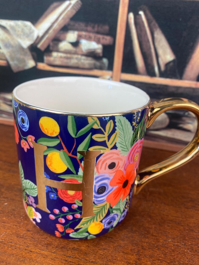 May include: A ceramic mug with a gold handle and the letter "H" in gold. The mug features a dark blue background with a floral pattern in various colors, including red, orange, and yellow. The inside of the mug is white.