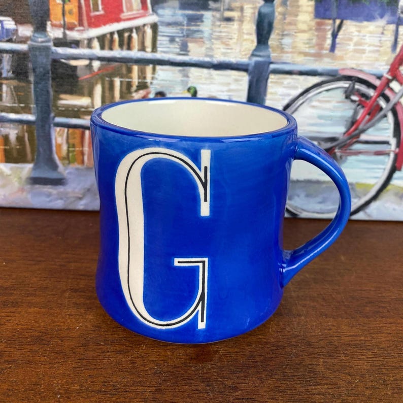 May include: A vibrant cobalt blue ceramic mug featuring a white interior and a prominent white letter "G". The mug, with its curved handle, is set against a backdrop of a painting with a bicycle.
