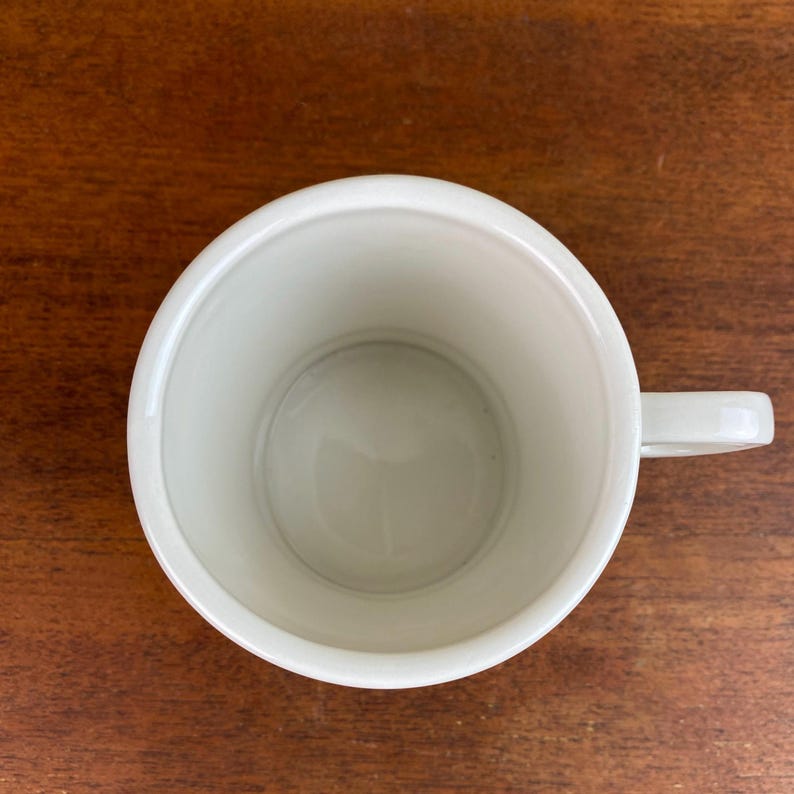 May include: A white ceramic mug with a rounded body and a small handle. The mug is empty and sits on a wooden surface. The interior of the mug is a light cream color.