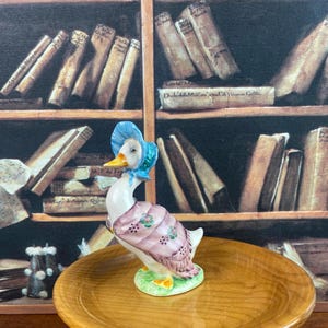 以下が含まれることがあります： 青い帽子とピンクのショールを着たガチョウの陶器製フィギュア。ガチョウは黄色の足が付いた緑色の台座の上に立っています。フィギュアは木製のプレートに飾られています。