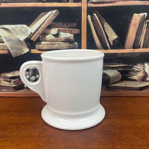 K&ouml;nnte beinhalten: Eine wei&szlig;e Keramiktasse mit rundem Boden und Henkel. Die Tasse hat ein schlichtes, klassisches Design, ideal f&uuml;r Kaffee oder Tee. Die Tasse steht vor einem Hintergrund aus B&uuml;chern.
