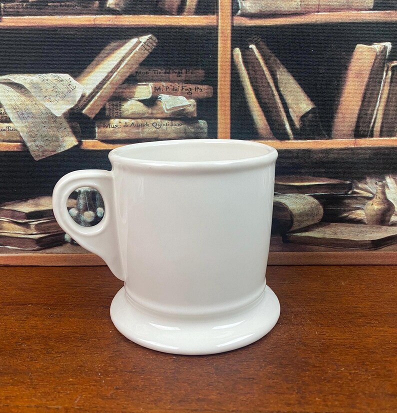 K&ouml;nnte beinhalten: Eine wei&szlig;e Keramiktasse mit rundem Boden und Henkel. Die Tasse hat ein schlichtes, klassisches Design, ideal f&uuml;r Kaffee oder Tee. Die Tasse steht vor einem Hintergrund aus B&uuml;chern.