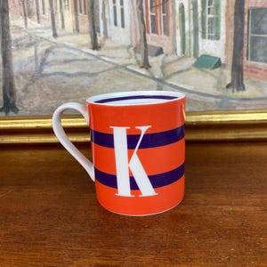 Può includere: Tazza in ceramica arancione con manico bianco, con una grande lettera bianca "K" e strisce orizzontali blu navy. La tazza ha un interno bianco e poggia su una superficie di legno.