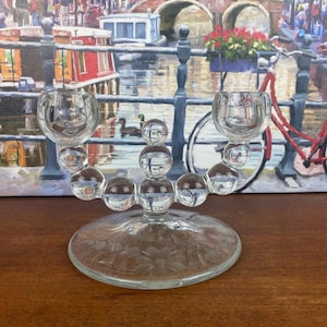 May include: Clear glass candelabra with two candle holders and a floral etched base. The candelabra features a series of clear glass spheres connecting the base to the candle holders. The background includes a painting of a canal scene.