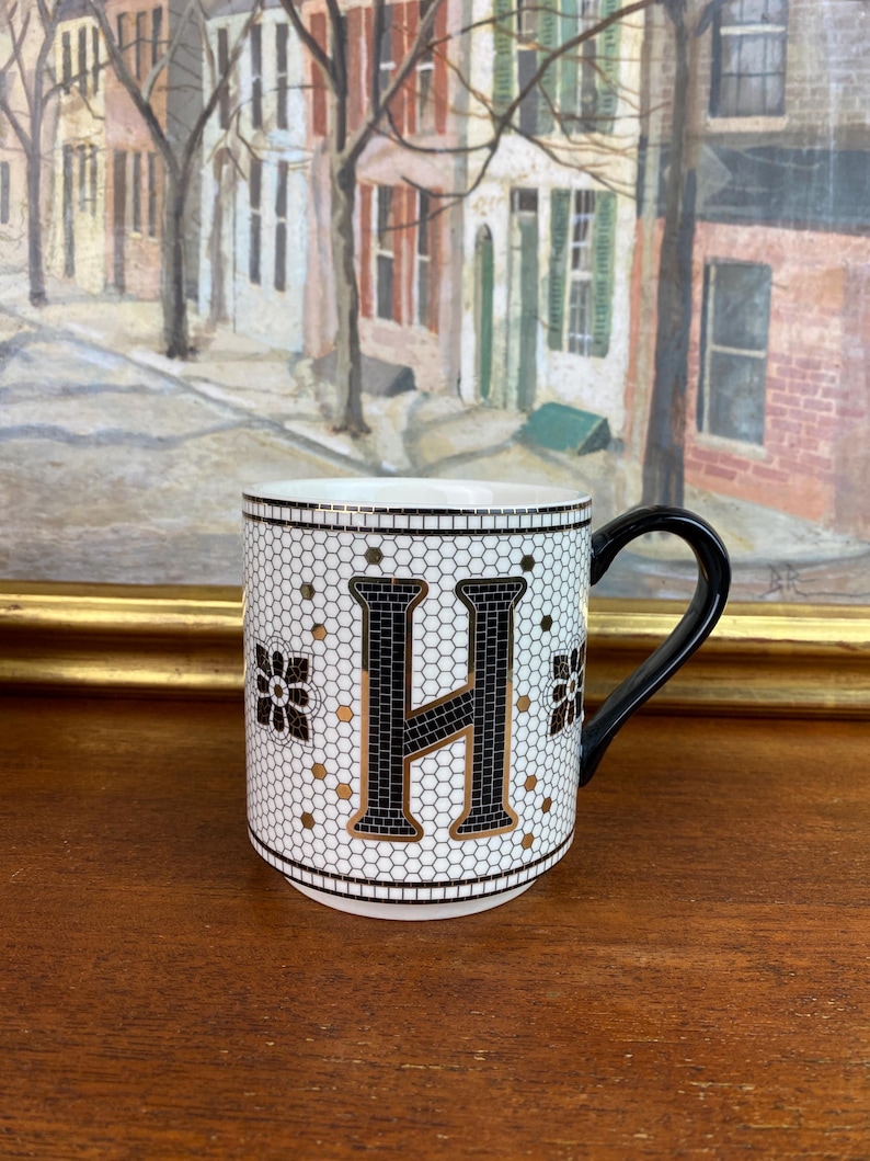 May include: White ceramic mug with a black and gold mosaic design featuring the letter "H".