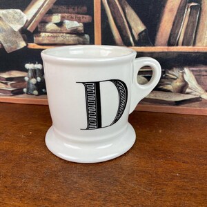 May include: White ceramic mug featuring a black letter "D". The mug has a rounded base and a handle. The background includes a bookshelf and books.