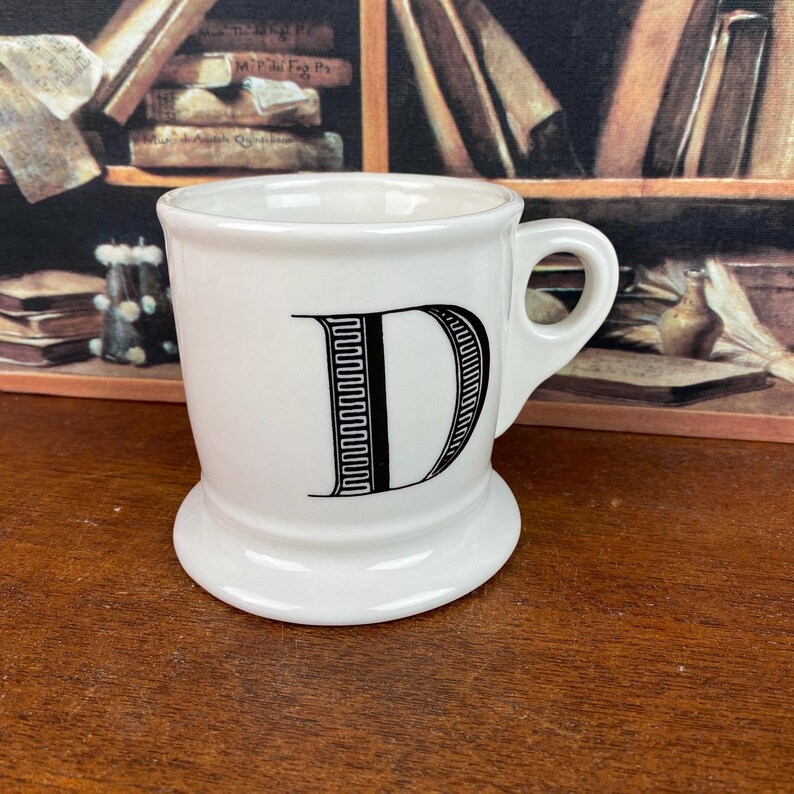 May include: White ceramic mug featuring a black letter "D". The mug has a rounded base and a handle. The background includes a bookshelf and books.