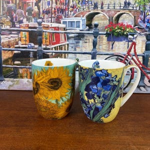 May include: Two ceramic mugs with floral designs. One mug features a large sunflower on a teal background. The other mug displays blue irises on a yellow background. Both mugs have white handles and rims, perfect for enjoying your favorite beverage.