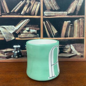 May include: A light green ceramic mug with a white interior and a large white letter "A" on the side. The mug is cylindrical and sits on a wooden surface. The background features a painting of a bookshelf.