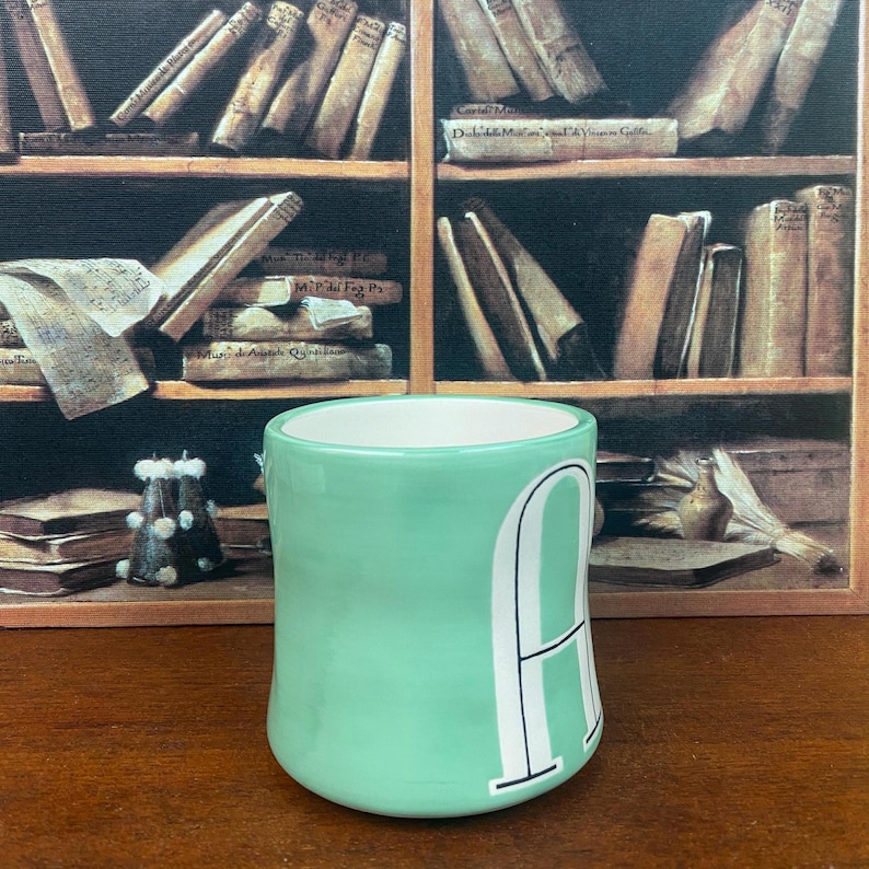 May include: A light green ceramic mug with a white interior and a large white letter "A" on the side. The mug is cylindrical and sits on a wooden surface. The background features a painting of a bookshelf.