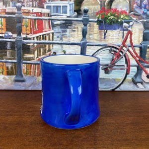 May include: A cobalt blue ceramic mug with a white interior and a curved handle. The mug has a glossy finish. The background features a painting of a canal scene with a red bicycle.
