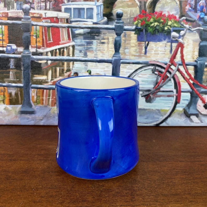 May include: A cobalt blue ceramic mug with a white interior and a curved handle. The mug has a glossy finish. The background features a painting of a canal scene with a red bicycle.