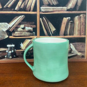 May include: A mint green ceramic mug with a curved handle. The interior of the mug is white. The mug is set against a backdrop of a bookshelf filled with books.
