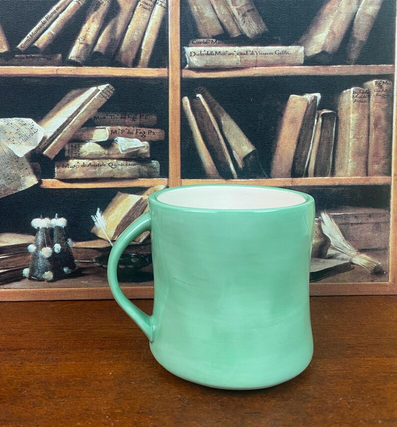 May include: A mint green ceramic mug with a curved handle. The interior of the mug is white. The mug is set against a backdrop of a bookshelf filled with books.