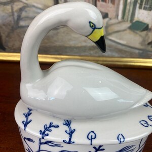 May include: A white ceramic swan figurine with a yellow beak and black tip, atop a round, lidded dish. The dish is white with blue aquatic plant and fish designs. The swan's neck is gracefully curved.