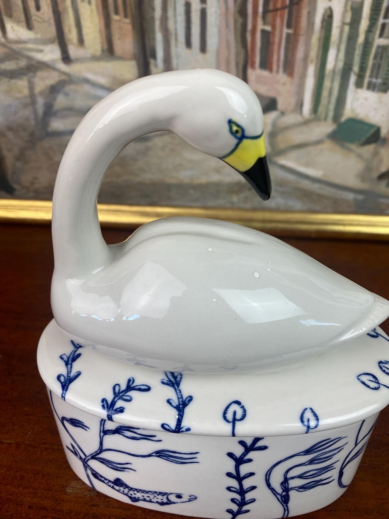 May include: A white ceramic swan figurine with a yellow beak and black tip, atop a round, lidded dish. The dish is white with blue aquatic plant and fish designs. The swan's neck is gracefully curved.
