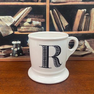 K&ouml;nnte beinhalten: Eine wei&szlig;e Keramiktasse mit einem gro&szlig;en schwarzen Buchstaben "R" auf der Vorderseite. Die Tasse hat einen abgerundeten Boden und einen Henkel. Der Hintergrund zeigt ein B&uuml;cherregal voller B&uuml;cher.