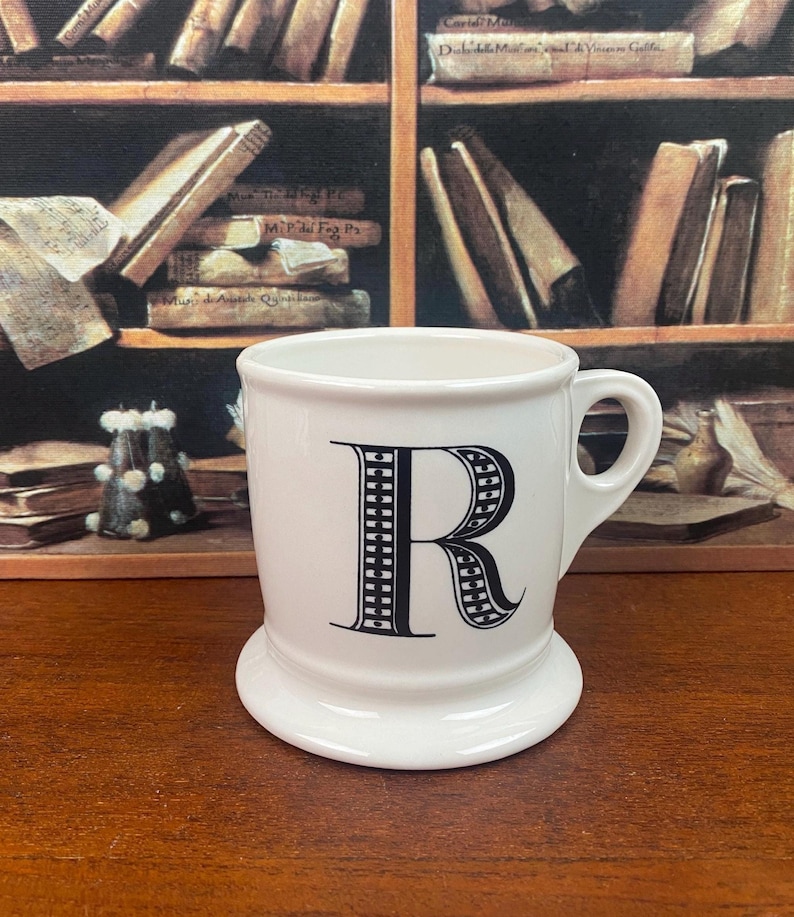 K&ouml;nnte beinhalten: Eine wei&szlig;e Keramiktasse mit einem gro&szlig;en schwarzen Buchstaben "R" auf der Vorderseite. Die Tasse hat einen abgerundeten Boden und einen Henkel. Der Hintergrund zeigt ein B&uuml;cherregal voller B&uuml;cher.