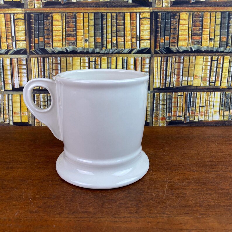 May include: A white ceramic shaving mug with a handle and a wide base. The mug has a classic design and is set against a backdrop of a bookshelf filled with books.