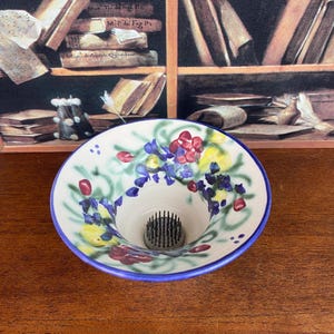 Puede incluir: Un jarrón de cerámica blanca con borde azul y forma acampanada. El jarrón está decorado con un estampado floral en rojo, amarillo y azul. El centro tiene una rejilla de alfileres metálicos.