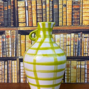 May include: A white ceramic vase with a green plaid design. The vase has a small handle and a rounded body. The background shows a bookshelf filled with books.