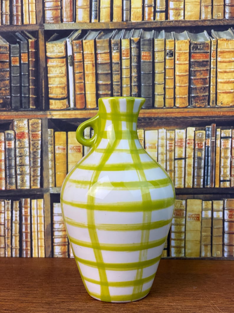May include: A white ceramic vase with a green plaid design. The vase has a small handle and a rounded body. The background shows a bookshelf filled with books.