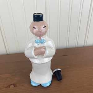 May include: A white ceramic figurine of a person wearing a blue and white robe and a black hat. The figurine is a bottle stopper with a silver top.