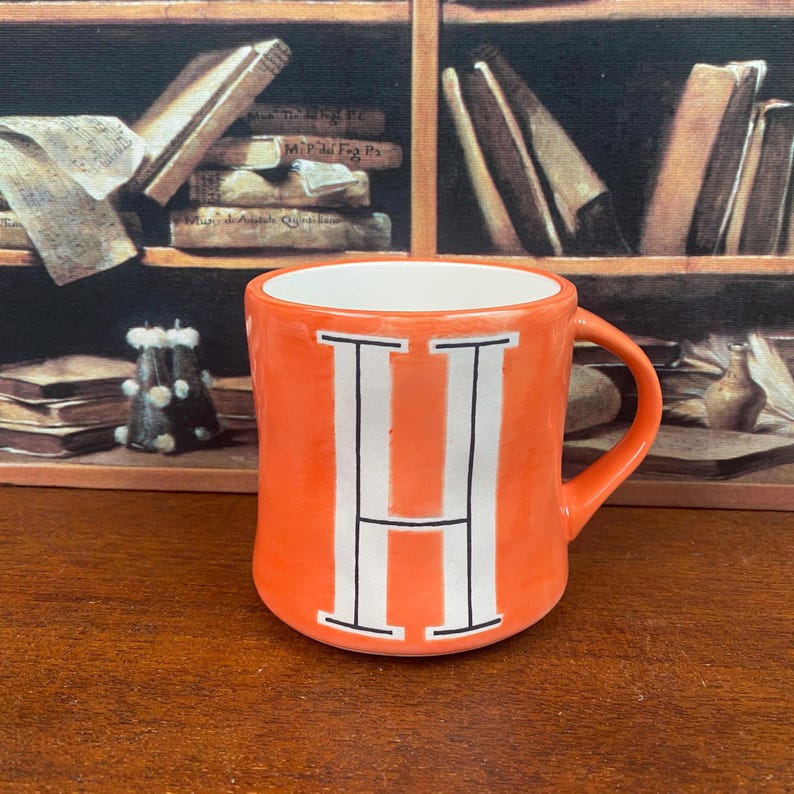 May include: Orange ceramic mug with a white interior and a large white letter "H" on the front. The mug has a curved handle and is set against a background of books and a wooden surface.