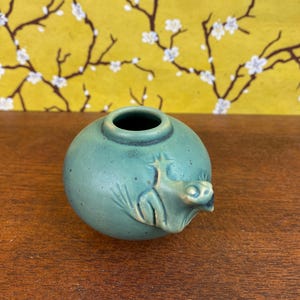 May include: A small, round, teal ceramic vase featuring a dragon sculpture. The vase has a dark rim and a small opening. The dragon has detailed features, including a snout and wings. The vase is a shade of teal.