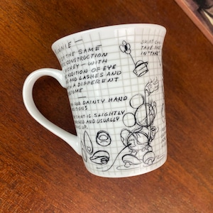 Puede incluir: Taza de cer&aacute;mica blanca con un boceto de Minnie Mouse, notas manuscritas y l&iacute;neas de cuadr&iacute;cula. La taza tiene un asa curva y un borde ligeramente acampanado. El dise&ntilde;o incluye texto y bocetos de los rasgos de Minnie.