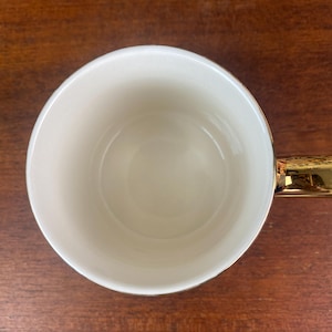 May include: A white ceramic mug with a gold handle and rim. The mug is empty and sits on a wooden surface. The interior of the mug is a creamy white color, and the handle is a shiny gold.