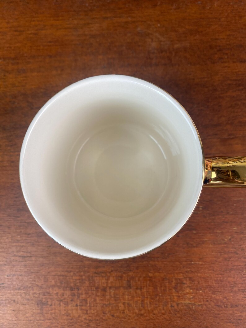 May include: A white ceramic mug with a gold handle and rim. The mug is empty and sits on a wooden surface. The interior of the mug is a creamy white color, and the handle is a shiny gold.
