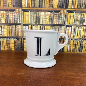 May include: White ceramic mug with a black letter "L" on the front. The mug has a rounded base and a handle. The background features a bookshelf filled with books.