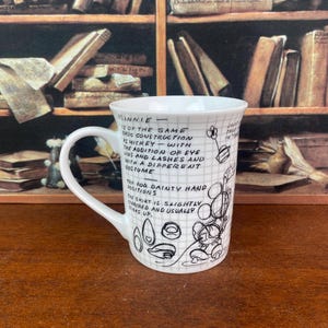 Puede incluir: Taza de cer&aacute;mica blanca con un patr&oacute;n de cuadr&iacute;cula negra y texto manuscrito y bocetos de Minnie Mouse. La taza tiene un asa curva y est&aacute; colocada sobre un fondo borroso de libros.