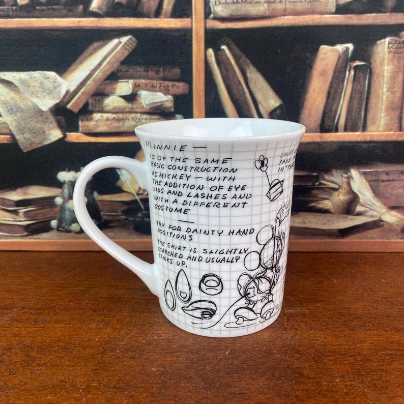 Puede incluir: Taza de cer&aacute;mica blanca con un patr&oacute;n de cuadr&iacute;cula negra y texto manuscrito y bocetos de Minnie Mouse. La taza tiene un asa curva y est&aacute; colocada sobre un fondo borroso de libros.