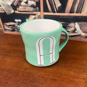 May include: A mint green ceramic mug with a white interior and a large white letter "A" on the front. The mug has a curved shape and a handle. The mug is sitting on a wooden surface.