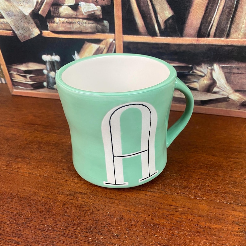 May include: A mint green ceramic mug with a white interior and a large white letter "A" on the front. The mug has a curved shape and a handle. The mug is sitting on a wooden surface.