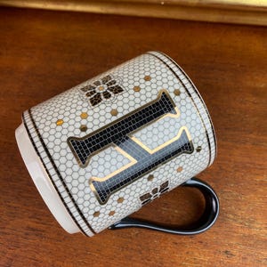 May include: White ceramic mug with a black and gold mosaic design featuring the letter "F". The mug has a black handle.
