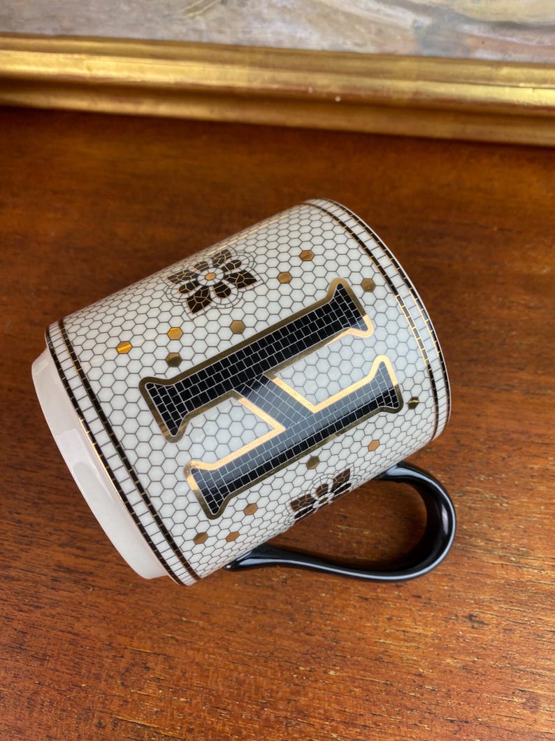 May include: White ceramic mug with a black and gold mosaic design featuring the letter "F". The mug has a black handle.