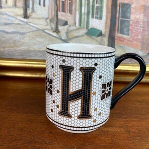 May include: White ceramic mug with a black letter H on a white and gold mosaic background. The mug has a black handle.