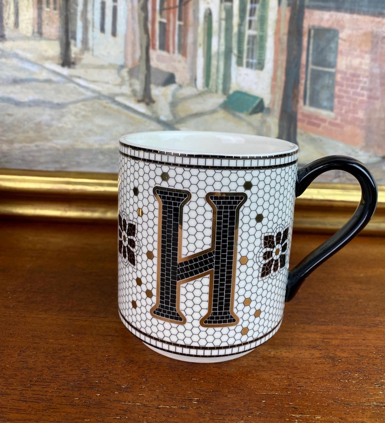 May include: White ceramic mug with a black letter H on a white and gold mosaic background. The mug has a black handle.
