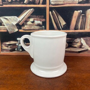 May include: A white ceramic mug with a handle and a wide, flat base. The mug has a simple, classic design and is likely used for drinking beverages. The background features a shelf with books.