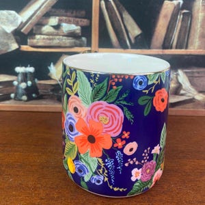 May include: A cylindrical ceramic mug with a navy blue background, adorned with a vibrant floral pattern in shades of pink, orange, blue, and green. The rim is white with a gold trim.