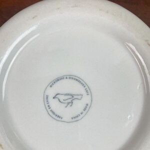 K&ouml;nnte beinhalten: Nahaufnahme des Bodens einer wei&szlig;en Keramiksch&uuml;ssel. Die Sch&uuml;ssel hat einen runden Stempel mit einer Vogelillustration und dem Text "MICROWAVE & DISHWASHER SAFE", "FABRIQUE EN CHINE" und "MADE IN CHINA".
