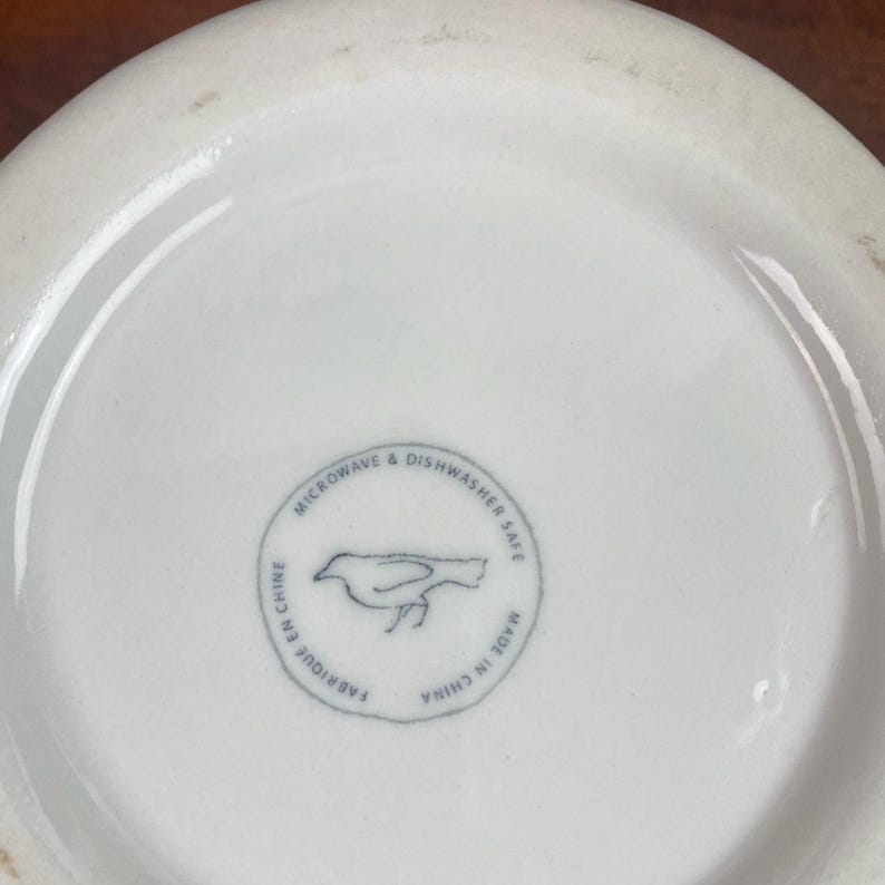 K&ouml;nnte beinhalten: Nahaufnahme des Bodens einer wei&szlig;en Keramiksch&uuml;ssel. Die Sch&uuml;ssel hat einen runden Stempel mit einer Vogelillustration und dem Text "MICROWAVE & DISHWASHER SAFE", "FABRIQUE EN CHINE" und "MADE IN CHINA".