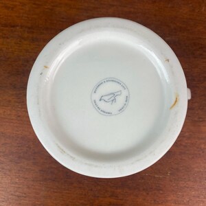 May include: White ceramic mug base with a small bird logo and text that reads "MICROWAVE & DISHWASHER SAFE" and "MADE IN CHINA". The mug is on a wooden surface.