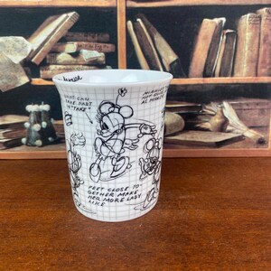 Puede incluir: Taza de cer&aacute;mica blanca con un boceto en blanco y negro de Minnie Mouse y texto manuscrito. La taza presenta un patr&oacute;n de cuadr&iacute;cula y varios bocetos de Minnie Mouse en diferentes poses. El texto incluye frases como "Shirt can take part in take" y "Feet close together make her more lady like."