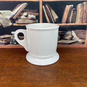 May include: A white ceramic shaving mug with a handle. The mug has a wide base and a rounded top. The handle is a loop shape. The mug is clean and simple, with a classic design.
